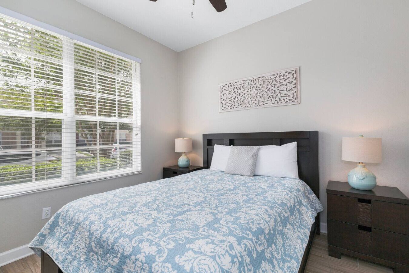 A bedroom with a bed, two bedside tables, a window with blinds, and a decorative wall piece.