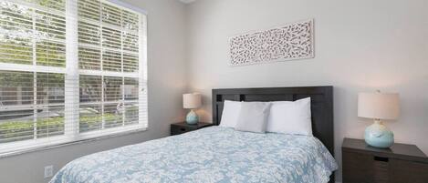 A bedroom with a bed, two bedside tables, a window with blinds, and a decorative wall piece.