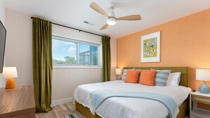 Habitación de hotel con cama, ventilador de techo, ventana con cortinas, mesita de noche con lámpara y un cuadro colgado en la pared.
