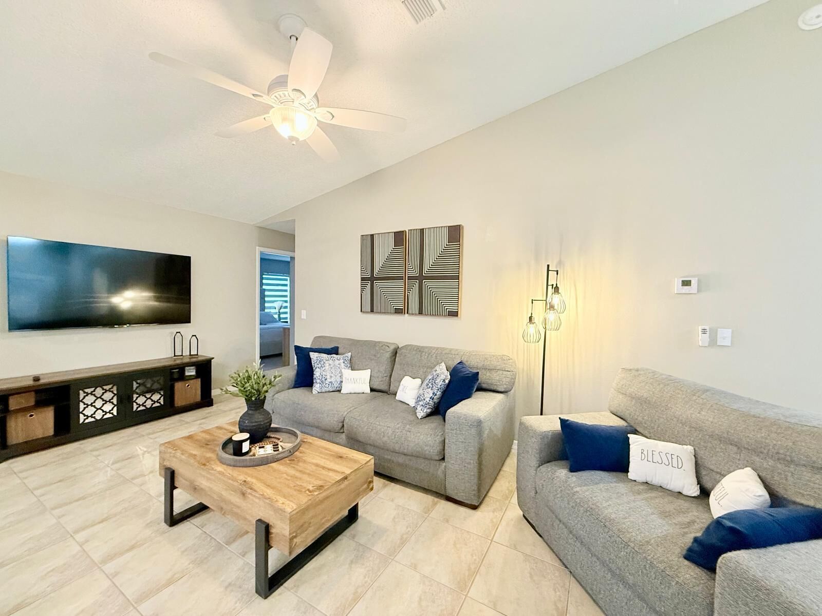 A modern living room with a flat-screen TV, grey sofas, a wooden coffee table, and a ceiling fan.