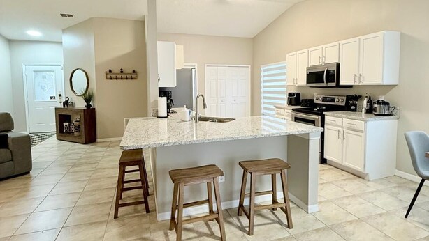 A modern kitchen with white cabinets, a central island with stools, and a dining area with a sofa and a coffee table. - Luxe & Modern Home-10 min to Disney Parks+Heated Pool+ Pets allowed (Kissimmee)