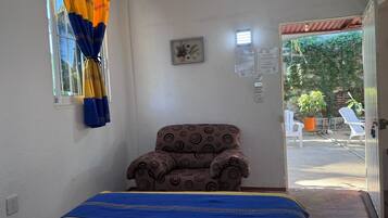 Una habitación con cama, un sillón estampado, una ventana con cortinas, un cuadro pequeño en la pared y una puerta que da al exterior.