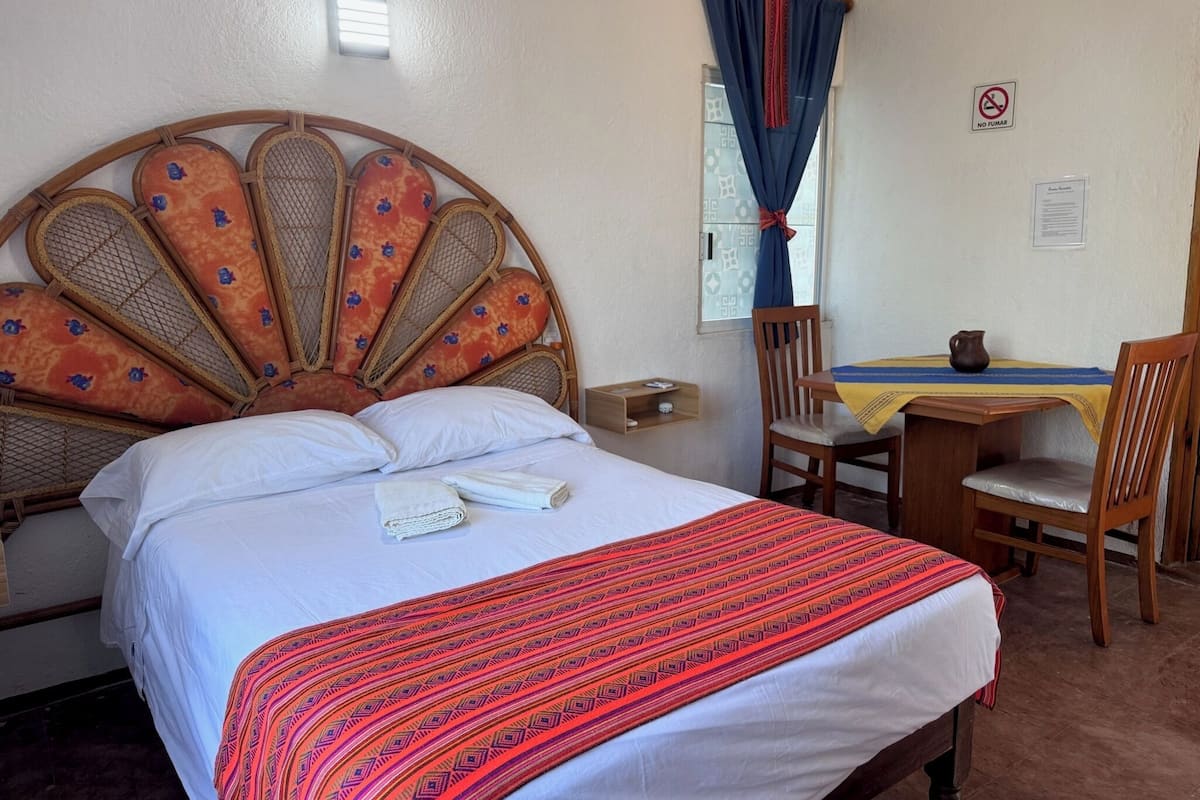 Habitación de hotel con cama, cabecera de madera, una mesita con jarrón y una silla.
