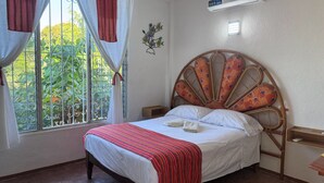 A bedroom with a bed, a window with curtains, a wall-mounted air conditioner, and a decorative wall piece. - Casa Icaco  (Puerto Escondido)