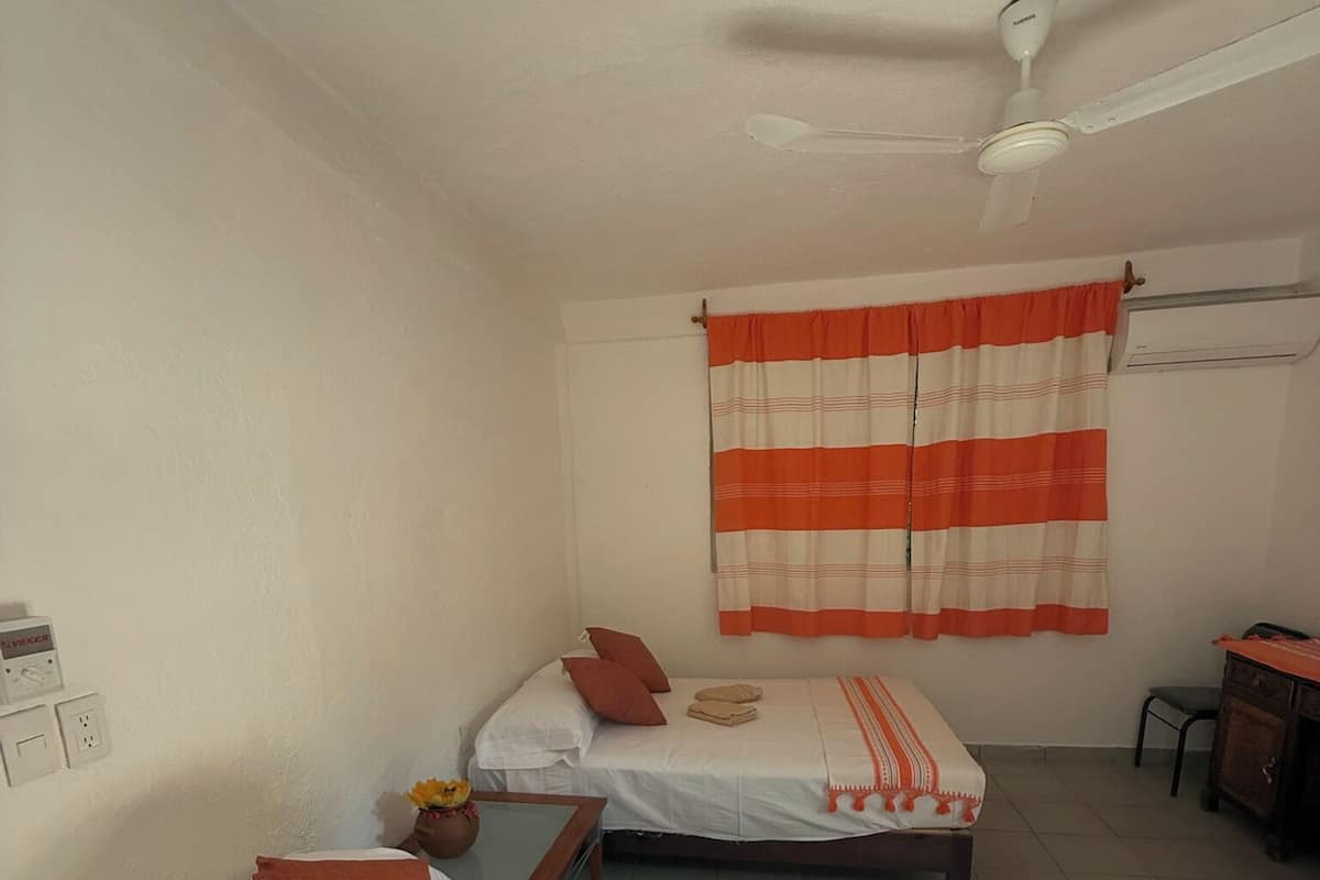 Una habitación con dos camas, un ventilador de techo, una ventana con cortina a rayas naranja y blanca, y una mesita con un florero.