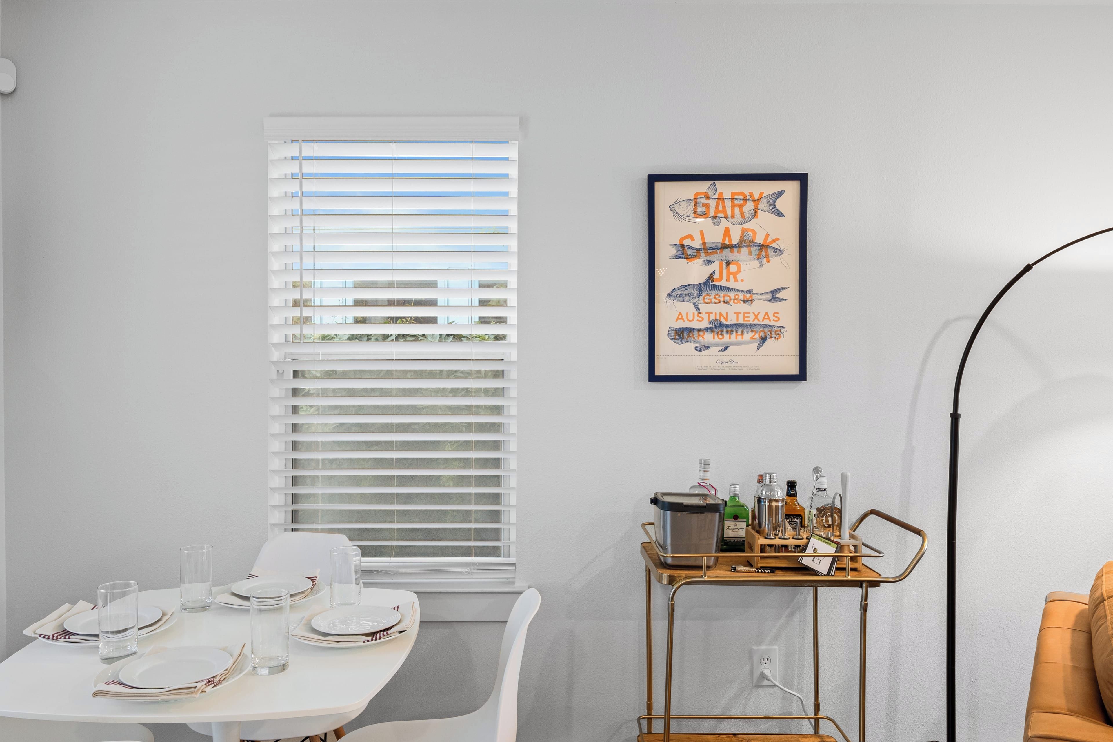 Un coin repas avec une table blanche dressée pour quatre personnes, une fenêtre avec des stores, une affiche encadrée au mur et un chariot rempli de bouteilles.
