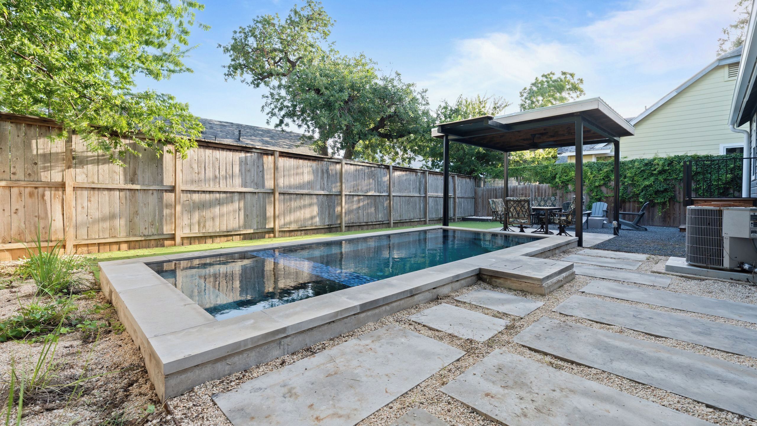Un jardin avec une piscine rectangulaire, une clôture en bois et un espace patio aménagé avec du mobilier d’extérieur.