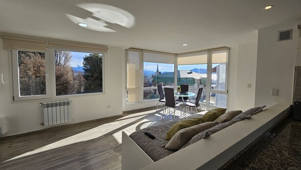 A modern living room with a sofa, a dining table with chairs, large windows, and a view of buildings and trees. - Andina View Bariloche (San Carlos de Bariloche)