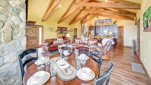 A spacious living room with a high ceiling, exposed wooden beams, a stone wall, and a dining area with a round table set for six. - Stunning Unobstructed Lake & Mountain Views, Hot Tub, In-Town, 1 mile to RMNP (Grand Lake)