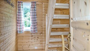 Interior de una cabaña de madera con ventana, cortinas de cuadros y una escalera de madera.