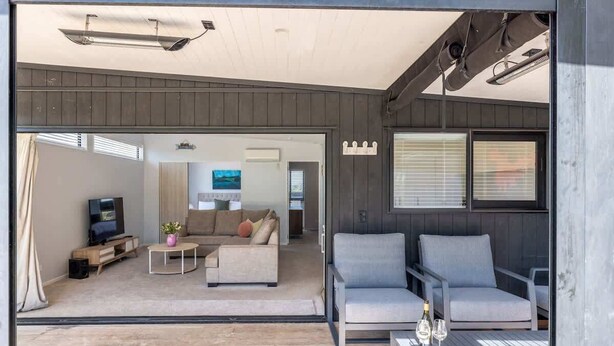 A modern living room with a sofa, armchairs, and a coffee table. There is a television and a small table with a plant. - Nikau Grove - Our Island (Waiheke Island)