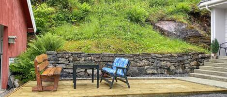 A wooden deck with two chairs and a table, a stone wall, and a red building in the background.