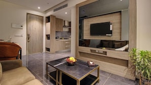 Una sala moderna con un sofá, una mesa baja con fruta, un televisor montado en la pared y una cocina visible a través de una puerta abierta.