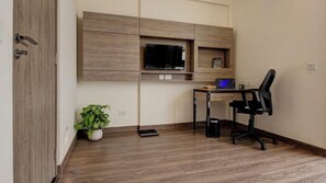 A room with a wooden floor, a desk with a laptop, a black office chair, a potted plant, and a mounted TV. - Rhythm Gurugram - Hotel & Suites (GURUGRAM)