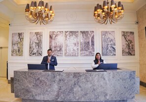 A reception desk with two individuals standing behind it, a chandelier above, and framed artwork on the wall. - Rhythm Gurugram - Hotel & Suites (GURUGRAM)