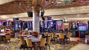 Interior de un casino con mesas de juego, sillas y máquinas tragamonedas.