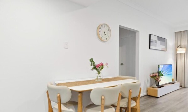 Un'area pranzo moderna con un tavolo rotondo, quattro sedie, un orologio, una porta, un televisore e un vaso con fiori.