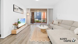A modern living room with a flat-screen TV, a sofa, a coffee table, and a view of a balcony with a brick building. - KozyGuru | Camperdown | Stylish Apt with Parking (Camperdown)