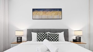 A hotel room with a large bed, two bedside tables with lamps, a checkered pillow, and a painting on the wall. - KozyGuru | Camperdown | Stylish Apt with Parking (Camperdown)
