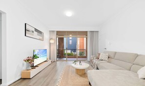 A modern living room with a flat-screen TV, a sofa, a coffee table, and a view of a balcony with a brick building.