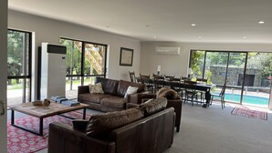 A spacious living room with a brown leather sofa, a dining table with chairs, and a large window overlooking a pool.