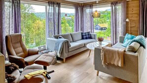 A living room with a sofa, armchair, and coffee table. There is a window with curtains, a lamp, and a view of greenery outside.