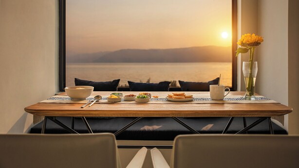 A dining table with a view of the sea at sunset, a vase with a single flower, and a glass of water.