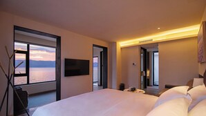 A modern hotel room with a large bed, a TV mounted on the wall, and a view of the sea through a large window.
