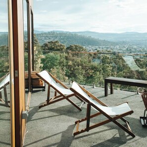 Un balcón con dos sillones de madera y una pequeña mesa, con vistas a un paisaje pintoresco de un valle y montañas lejanas.
