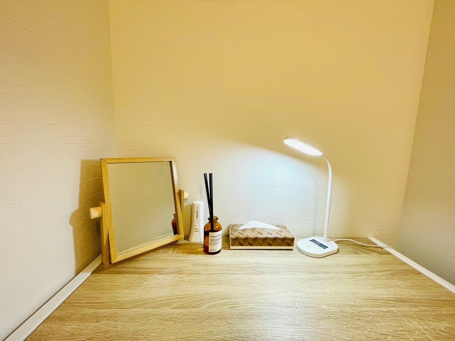 Um quarto pequeno com piso de madeira, parede branca, espelho emoldurado, abajur, frasco de álcool em gel e caixa de lenços.