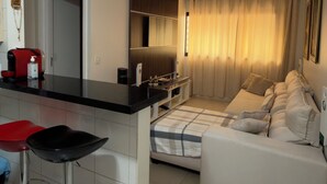 Una cocina moderna con encimera negra y taburetes de acero inoxidable, una zona de comedor con sofá blanco y una ventana con cortinas, y un pasillo que conduce a otra habitación.