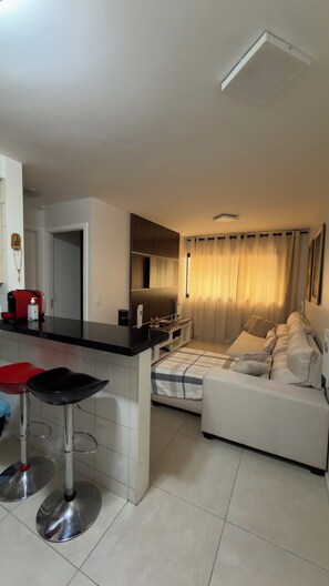 A modern kitchen with a black countertop and stainless steel stools, a dining area with a white sofa and a window with curtains, and a hallway leading to another room.
