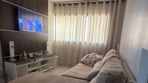 Ett modernt vardagsrum med en stor beige soffa, en platt-TV som är monterad på en träpanel, en liten vit TV-möbel och ett fönster med tunna, genomskinliga gardiner.