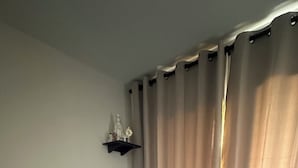 Una cama doble con una colcha color beige, dos toallas enrolladas, un estante con adornos y una ventana con cortinas.