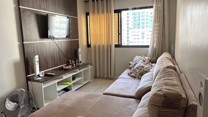 Ett modernt vardagsrum med en stor hörnsoffa, en platt-TV som är monterad på en träpanel, en liten TV-möbel och ett stort fönster med gardiner.