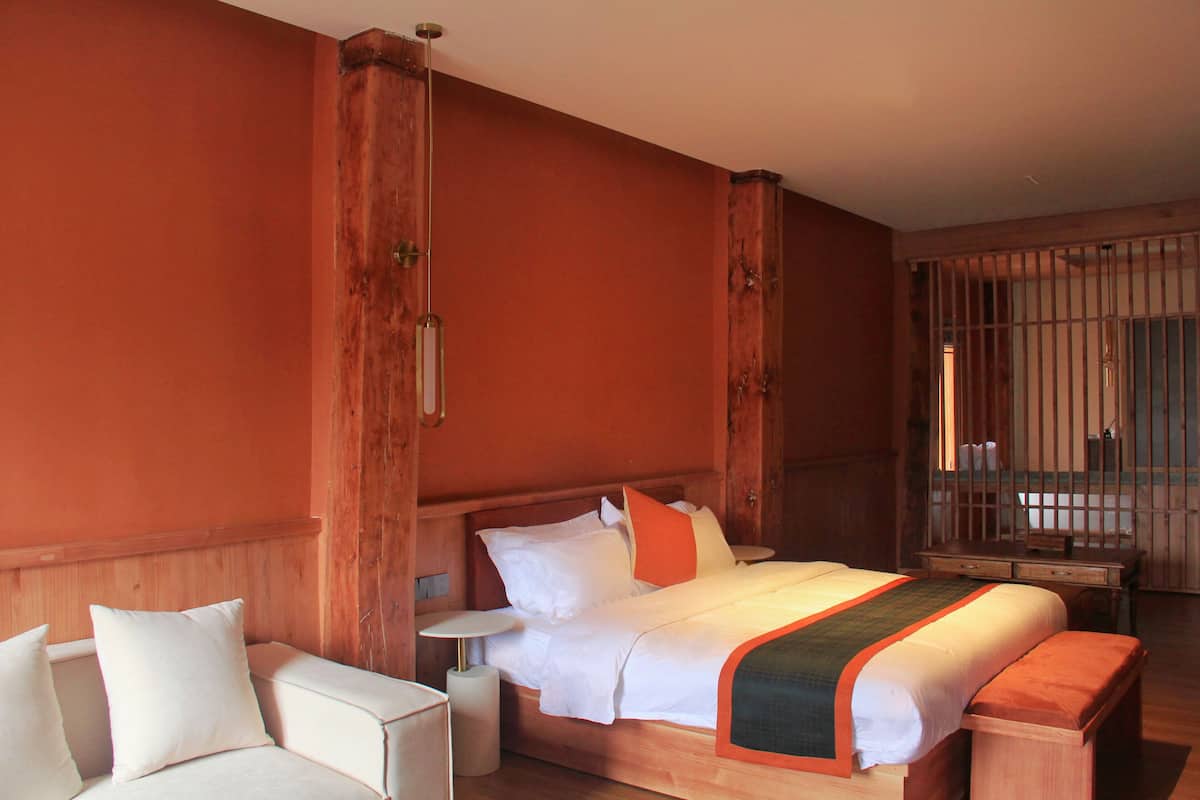 Habitación de hotel con cabecera de madera, cama con ropa de cama blanca y detalles naranja, mesita de noche y una silla.