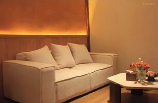 Un sofá color beige con dos almohadas, una mesita con un florero y un panel de madera detrás del sof á.