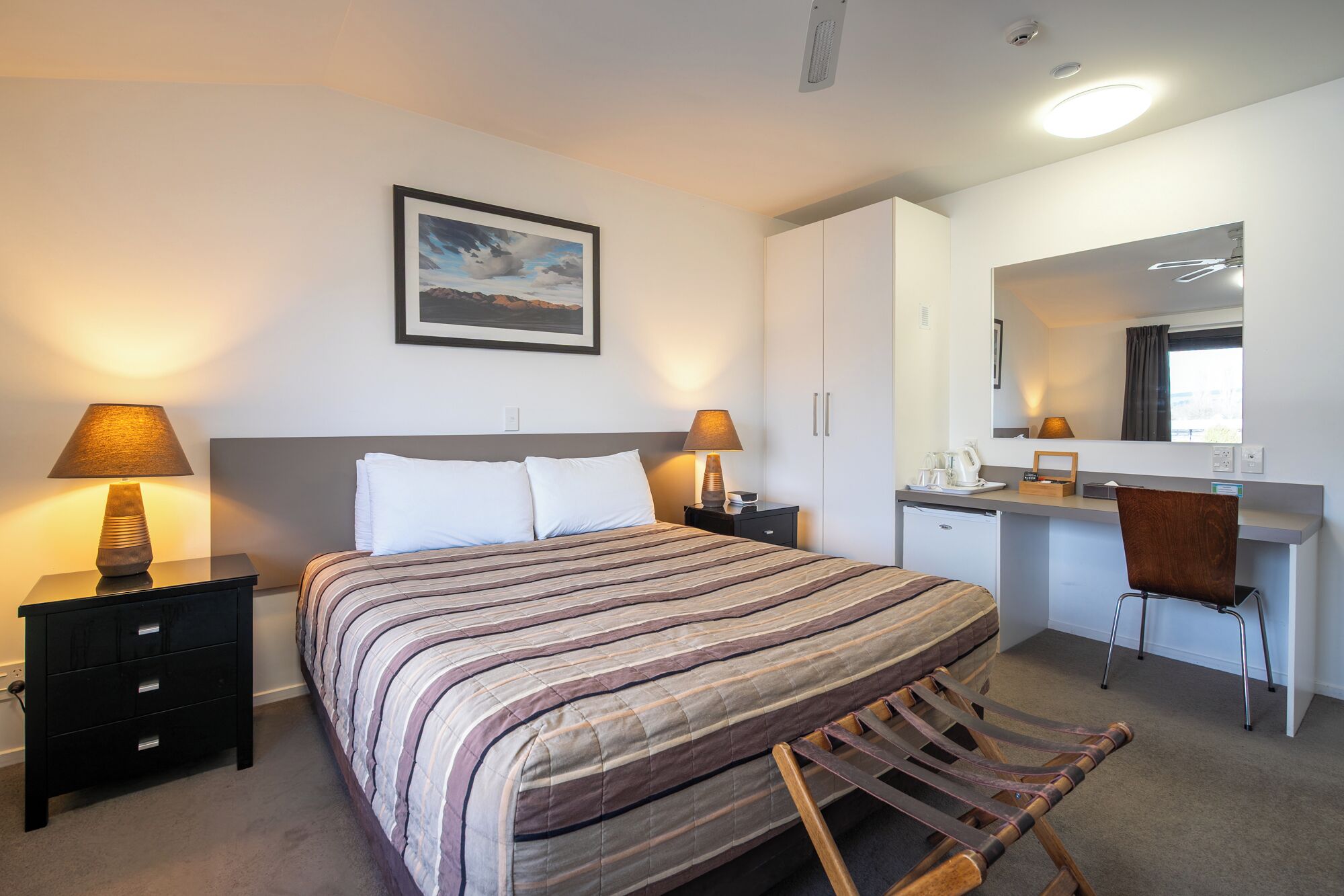 A hotel room with a bed, bedside tables, a desk, a chair, a mirror, and a framed picture on the wall.