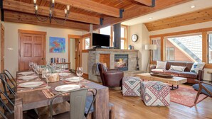 A spacious living area with a fireplace, wooden ceiling beams, and a dining table set for six. - Ski In Ski Out Big Sky Chalet (Big Sky)