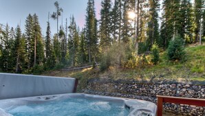 A hot tub with a stone wall and a wooden deck, surrounded by a forest and a rocky hillside. - Ski In Ski Out Big Sky Chalet (Big Sky)