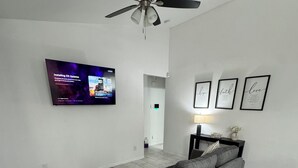 A modern living room with a grey sectional sofa, a ceiling fan, a flat-screen TV mounted on the wall, and a hallway leading to another room. - Perfect for Families & Groups in Fortuna Foothills (Yuma)