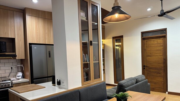 Une cuisine moderne avec des meubles en bois, un réfrigérateur encastré, un coin repas avec une table en bois et des sièges recouverts d’un tissu gris, et un ventilateur de plafond.