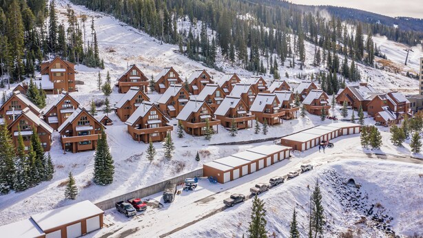 Una cabaña cubierta de nieve con varias cabañas de madera y un edificio central, rodeada de árboles y una pista de esquí.