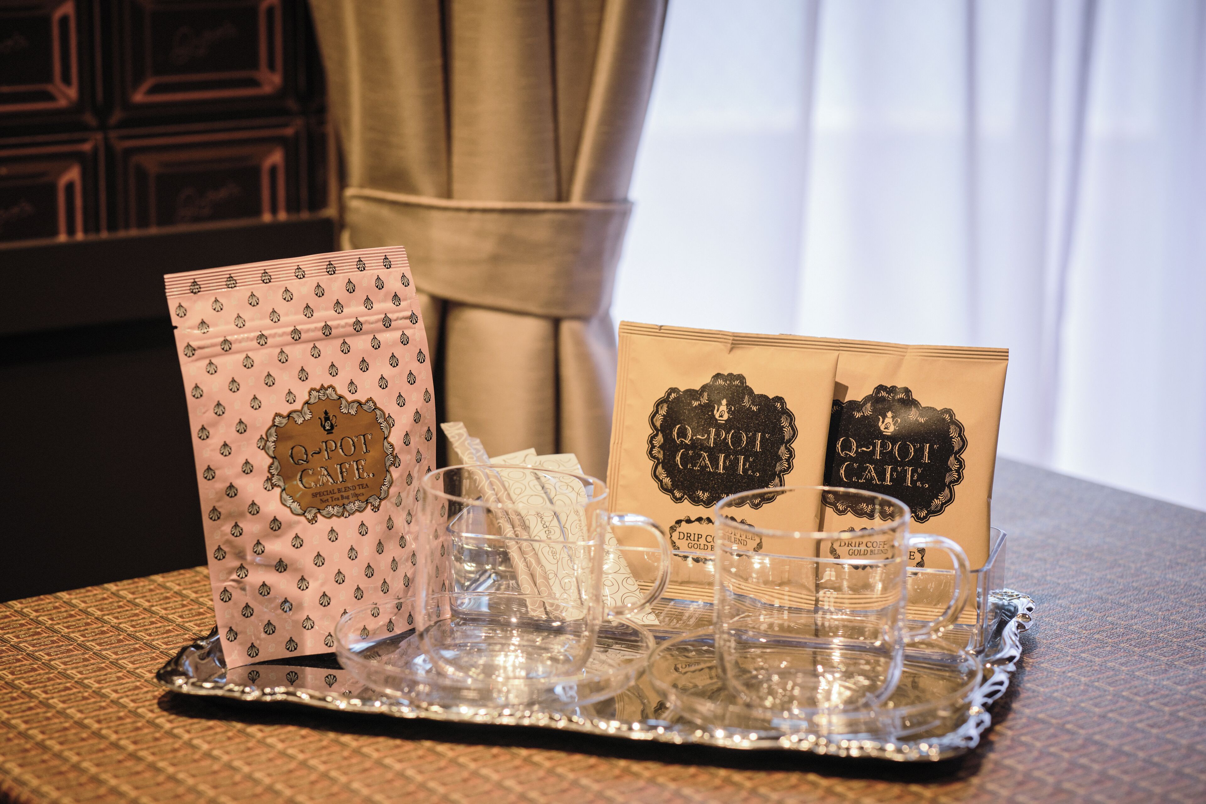 A tray with clear glass cups and saucers, two Q-pot cafe branded bags, and a patterned tablecloth.