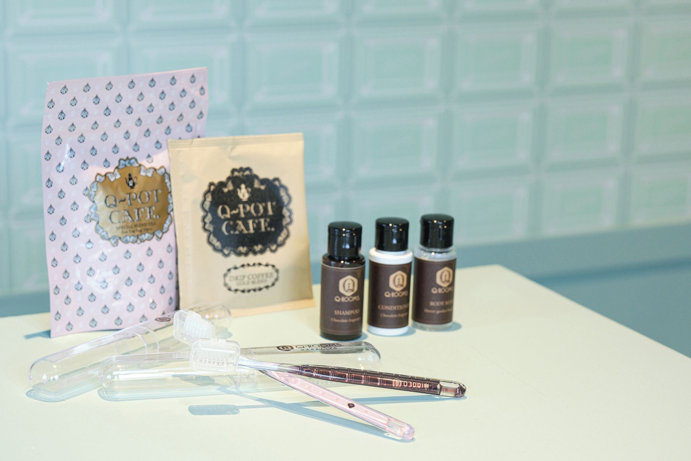 A bathroom vanity with a white countertop, a pink toothbrush holder, a Q-pot cafe branded bag, and four Q-pot cafe hair care products.