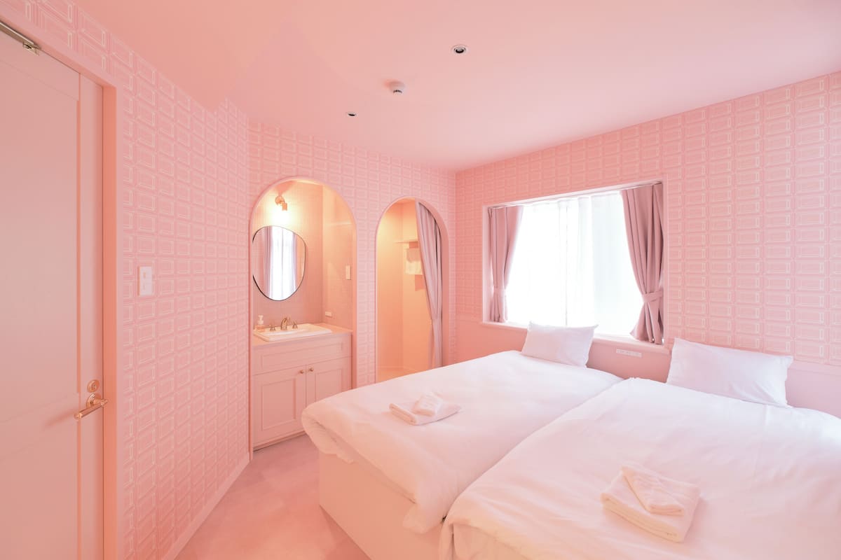 A pink-themed hotel room with a double bed, a vanity with a mirror, a window with curtains, and a door.