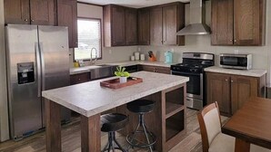 A modern kitchen with wooden cabinets, stainless steel appliances, a central island with stools, and a window with blinds.