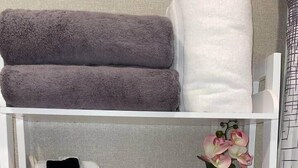 A bathroom shelf with towels, a woven basket, a vase with flowers, and a container with toiletries.