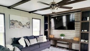A modern living room with a grey sofa, a large flat-screen TV, wooden shelves, and a ceiling fan.