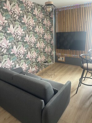 A modern living room with a grey sofa, a wooden slatted wall, and a large flat-screen TV. - 1-bedroom apartment With sofa bed 140 Fully equipped Functional Flexible (Favières)